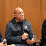 Three individuals sit together in a welcoming indoor space at Temple Emanu-El; two men are holding microphones engaging in conversation, while a woman attentively listens.