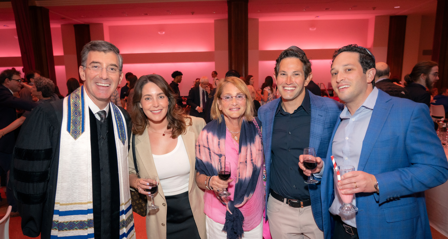Five people smile at a formal Temple Emanu-El event, holding beverages, with pink lighting in the background.