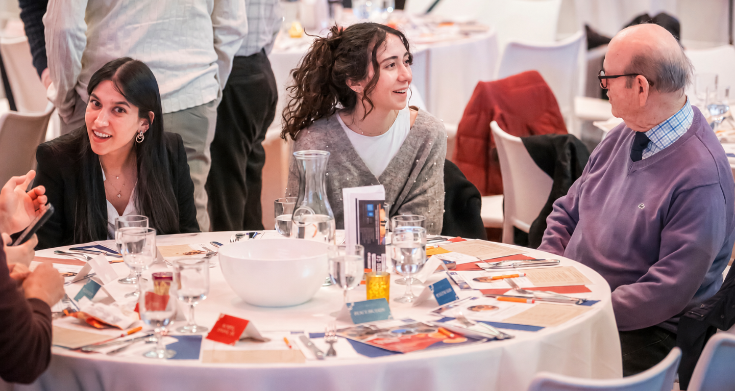 Three people sit at a round table set for a meal, engaging in warm conversation and smiling in a bright gathering space at Temple Emanu-El.