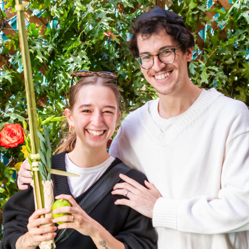 Two congregants smiling in front of lush greenery, one holding a lulav and etrog in celebration of Sukkot at Temple Emanu-El.
