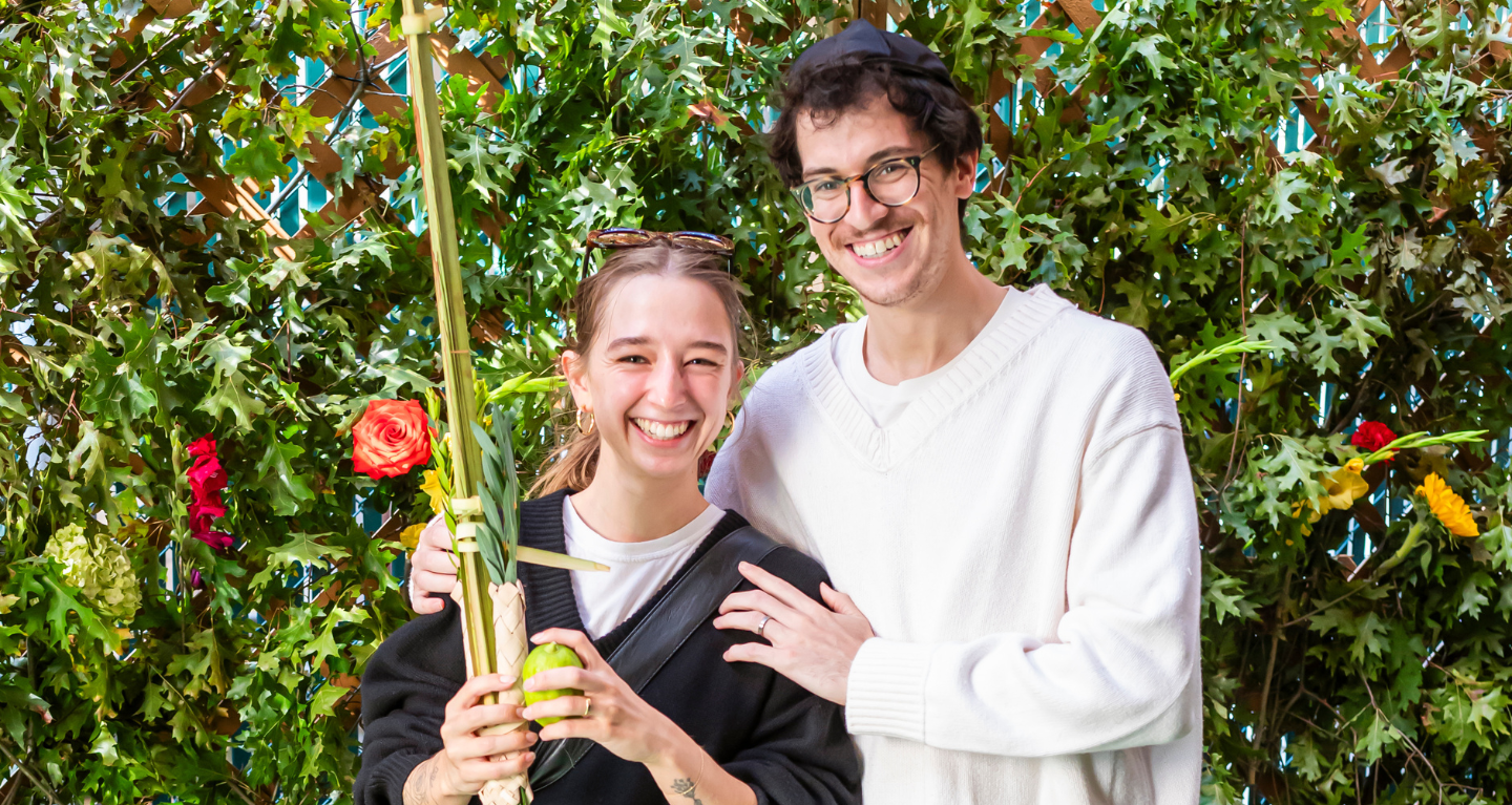 Two congregants smiling in front of lush greenery, one holding a lulav and etrog in celebration of Sukkot at Temple Emanu-El.