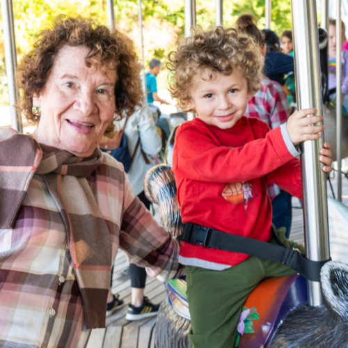 An older woman and a young child share joyful smiles while riding a carousel together on a sunny day, celebrating special moments of community and connection.