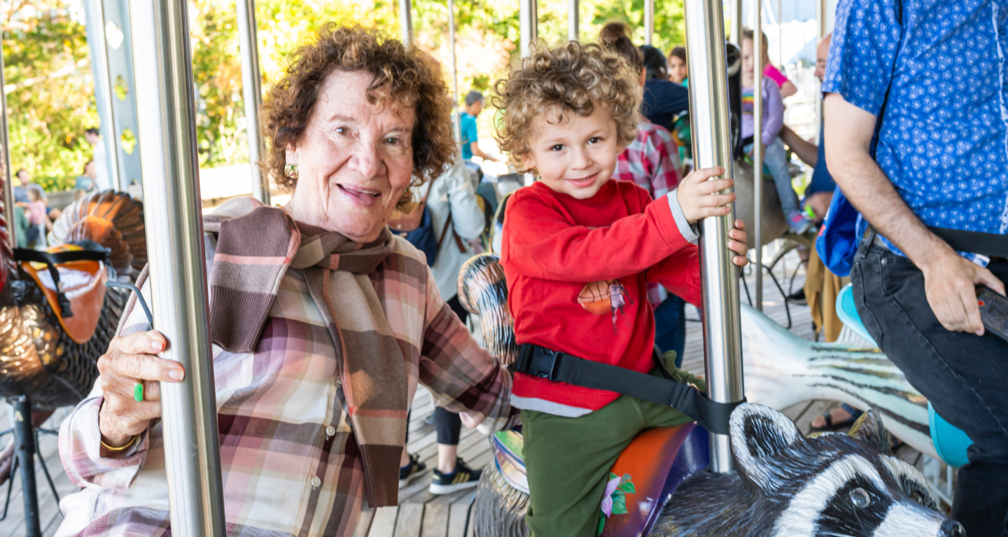 An older woman and a young child share joyful smiles while riding a carousel together on a sunny day, celebrating special moments of community and connection.