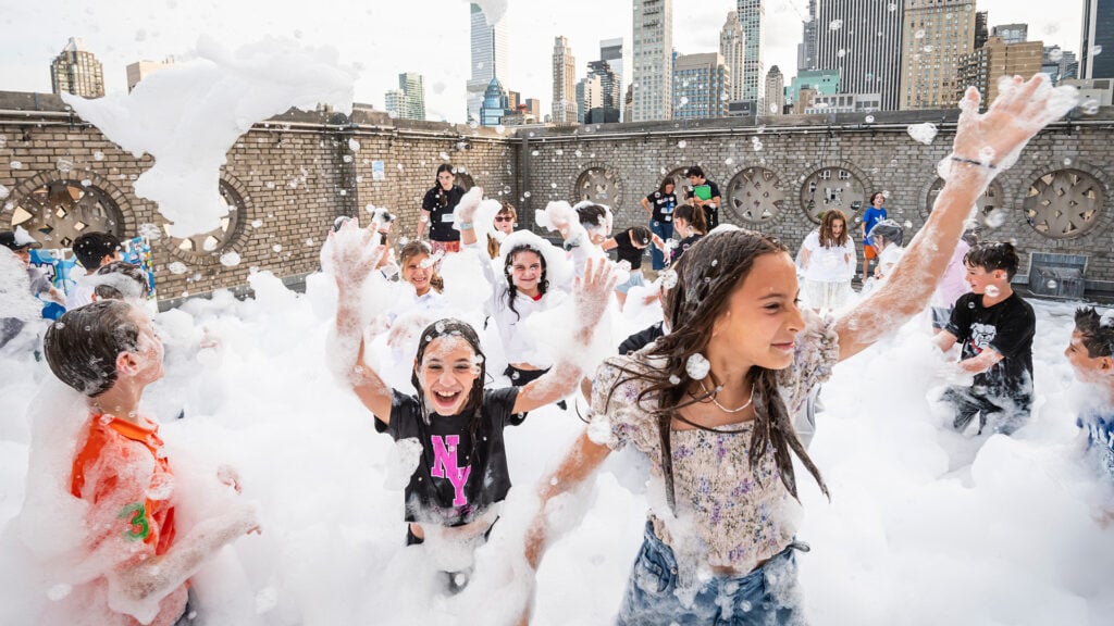 Children play and laugh in thick foam during a joyful gathering on a rooftop, with the iconic New York City skyline visible behind Temple Emanu-El.
