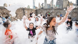 Children play and laugh in thick foam during a joyful gathering on a rooftop, with the iconic New York City skyline visible behind Temple Emanu-El.