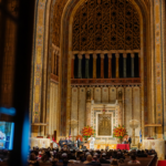 A large audience gathers for an event in the majestic sanctuary of Temple Emanu-El, surrounded by tall arches and elegant floral arrangements.