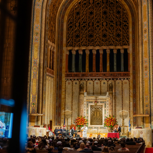 A large audience gathers for an event in the majestic sanctuary of Temple Emanu-El, surrounded by tall arches and elegant floral arrangements.