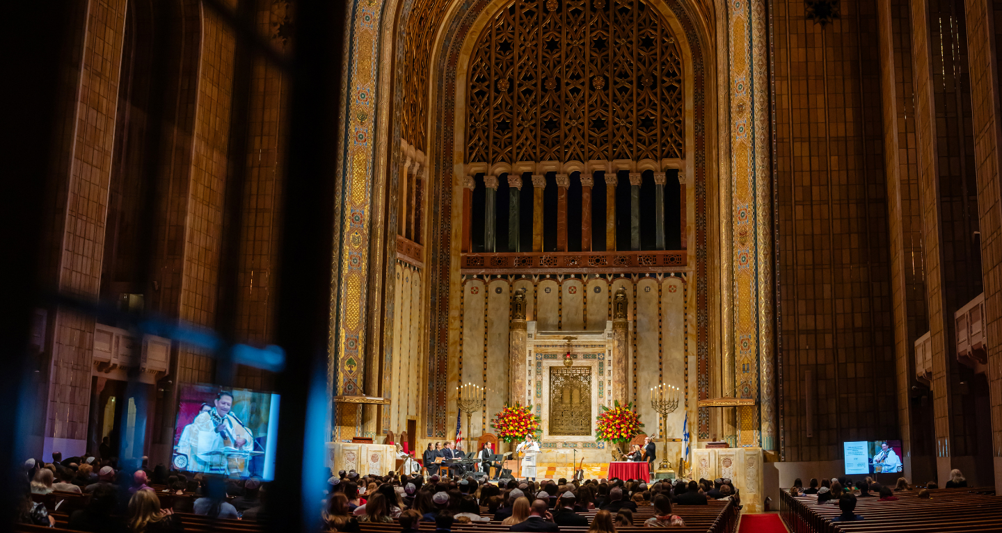 A large audience gathers for an event in the majestic sanctuary of Temple Emanu-El, surrounded by tall arches and elegant floral arrangements.