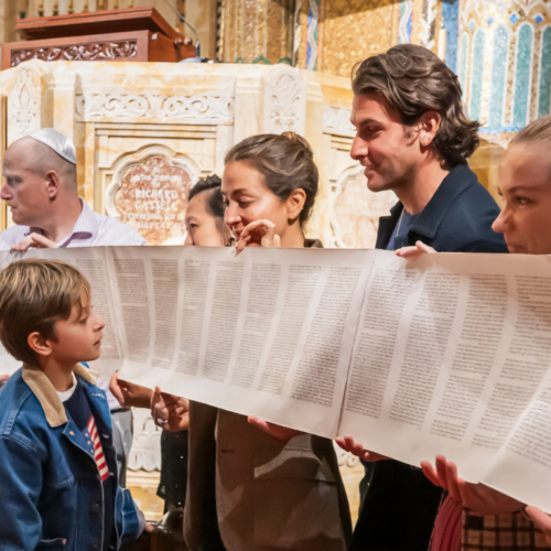Congregants of all ages, including children, hold a Torah scroll with Hebrew text inside the beautifully adorned sanctuary at Temple Emanu-El.