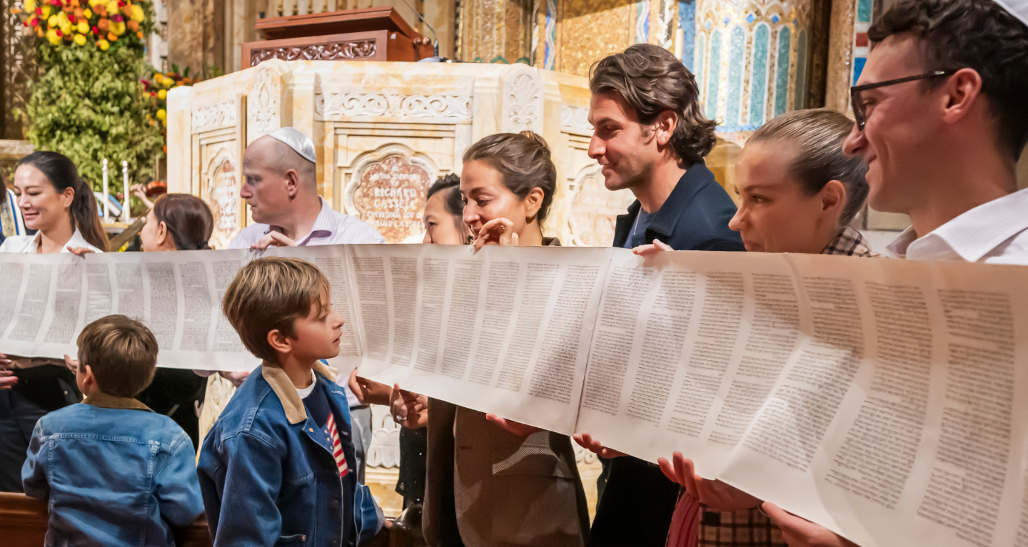 Congregants of all ages, including children, hold a Torah scroll with Hebrew text inside the beautifully adorned sanctuary at Temple Emanu-El.