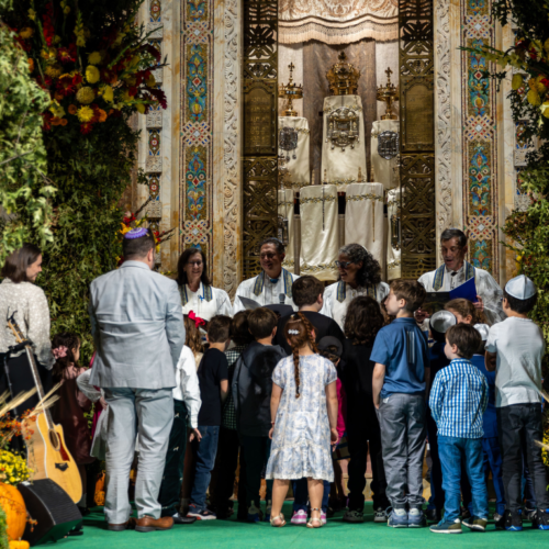 Children and adults gather around a beautifully decorated bimah with fruits, flowers, and greenery during a festive celebration at Temple Emanu-El.