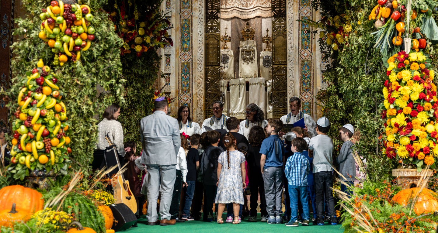 Children and adults gather around a beautifully decorated bimah with fruits, flowers, and greenery during a festive celebration at Temple Emanu-El.