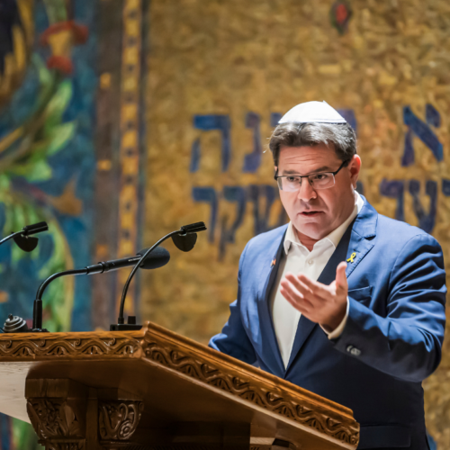 Man in suit and kippah speaks at an elegant podium in front of a decorative wall featuring Hebrew inscriptions at Temple Emanu-El.