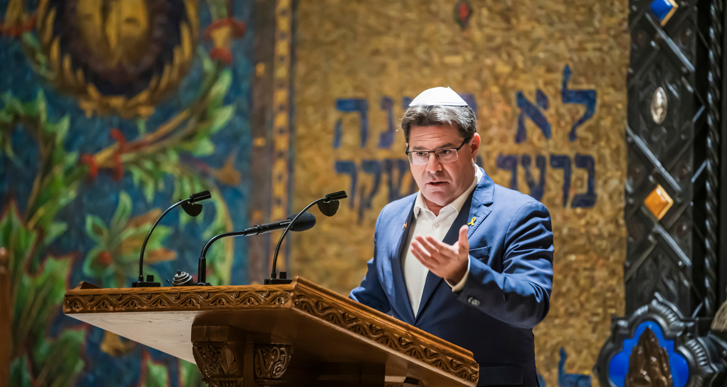 Man in suit and kippah speaks at an elegant podium in front of a decorative wall featuring Hebrew inscriptions at Temple Emanu-El.