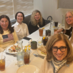 Seven women gather around a table at a Temple Emanu-El event, enjoying food and drinks together and smiling at the camera.