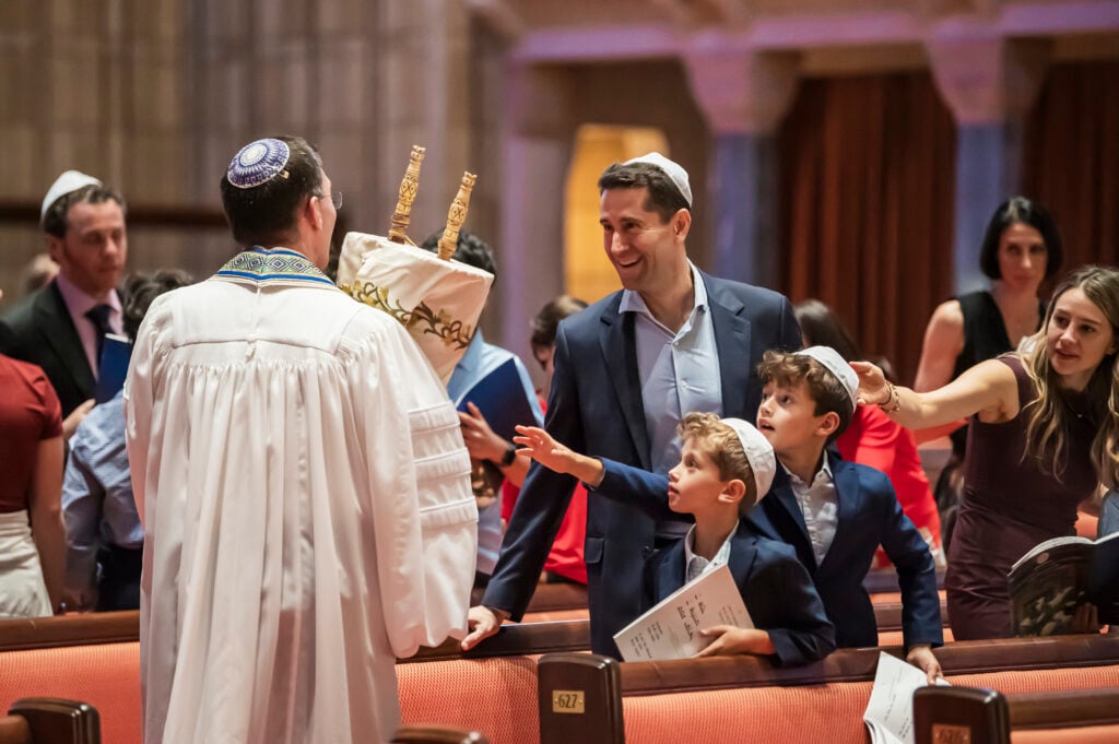 A Temple Emanu-El rabbi holding a Torah scroll warmly welcomes a smiling family with children inside the sanctuary.