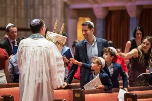 A Temple Emanu-El rabbi holding a Torah scroll warmly welcomes a smiling family with children inside the sanctuary.