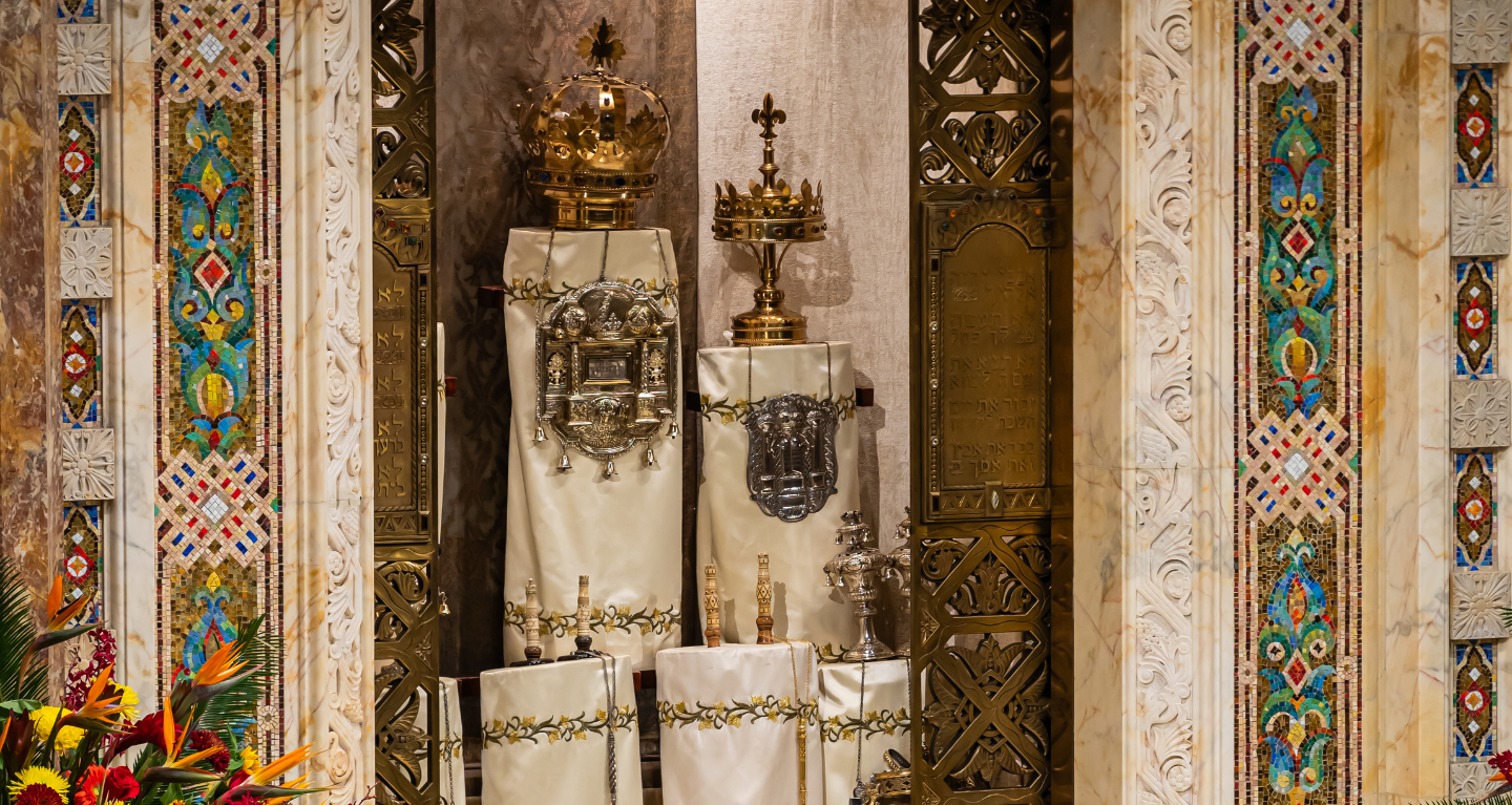 Torah scrolls adorned with elegant covers and decorative crowns displayed in a beautifully designed ark featuring detailed floral motifs at Temple Emanu-El.