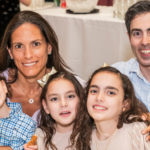 A family of four, two adults and two children, share a warm smile together at a table inside Temple Emanu-El’s bright communal space.