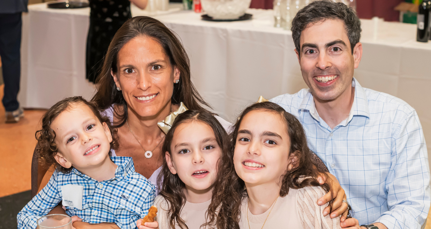 A family of four, two adults and two children, share a warm smile together at a table inside Temple Emanu-El’s bright communal space.