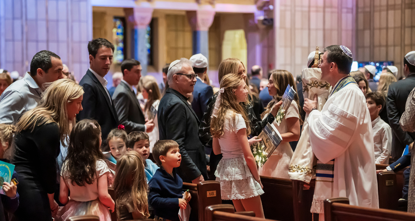 A rabbi welcomes children and adults holding prayer books in the sanctuary at Temple Emanu-El, with sunlight filtering through the ornate stained glass windows.