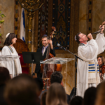 Four musicians dressed in white robes perform on stage at Temple Emanu-El; one musician sounds a shofar as the audience looks on.