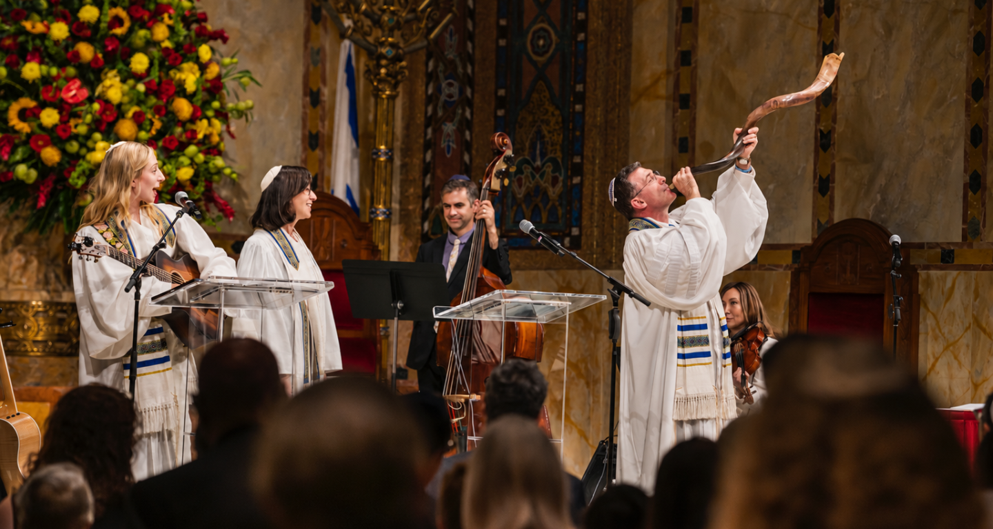 Four musicians dressed in white robes perform on stage at Temple Emanu-El; one musician sounds a shofar as the audience looks on.