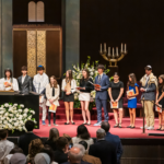 A group of young people stand on the bimah at Temple Emanu-El during a ceremony, proudly holding certificates.