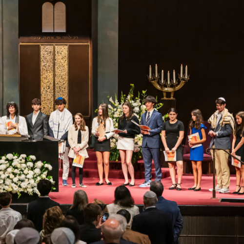 A group of young people stand on the bimah at Temple Emanu-El during a ceremony, proudly holding certificates.