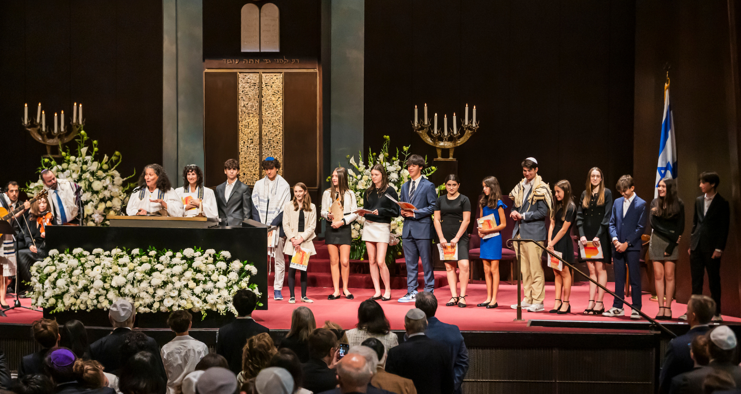 A group of young people stand on the bimah at Temple Emanu-El during a ceremony, proudly holding certificates.
