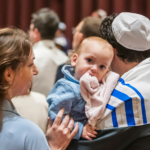 A baby in a blue outfit is held by an adult during a lively gathering at Temple Emanu-El, with community members present in the background.