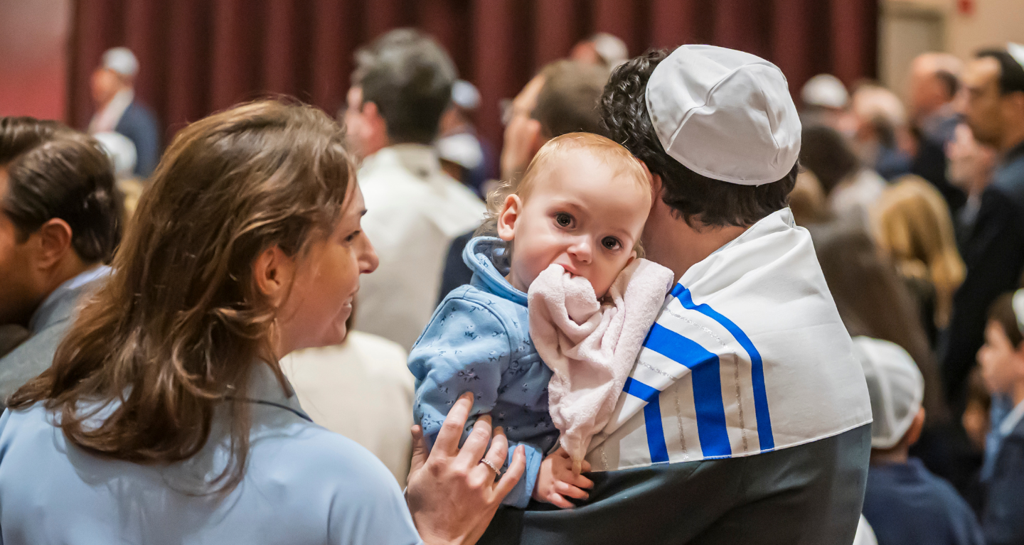 A baby in a blue outfit is held by an adult during a lively gathering at Temple Emanu-El, with community members present in the background.