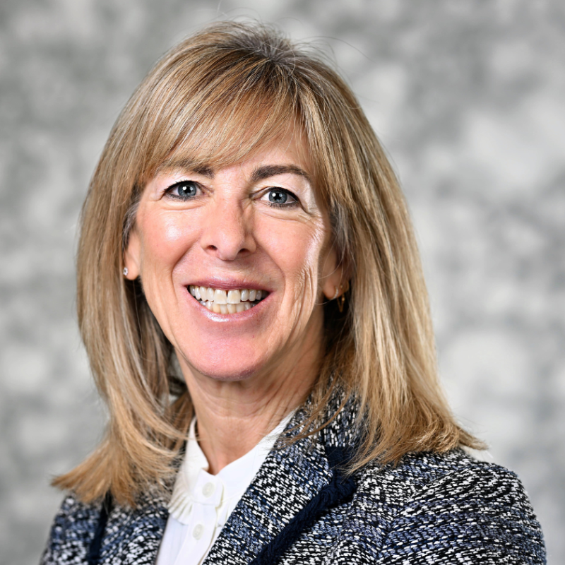 Smiling woman with blonde hair in a tweed jacket and white blouse, standing before a soft gray background at Temple Emanu-El.
