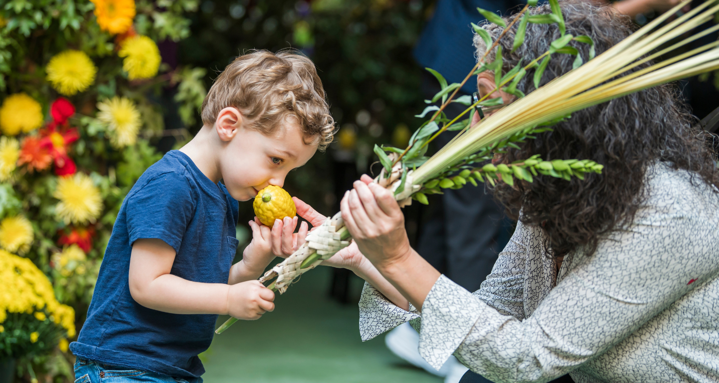 A child enjoys the fragrance of an etrog, gently held by an adult, during a Sukkot celebration at Temple Emanu-El with festive flowers adorning the background.