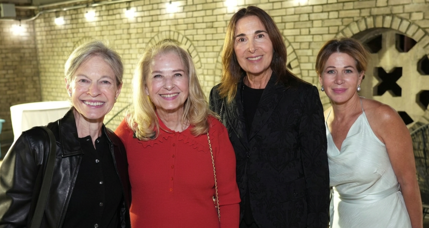 Four women smiling together at a Temple Emanu-El event, standing indoors in front of a brick wall adorned with lights.