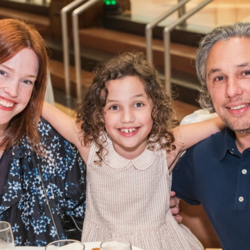 Three people smiling together at a table inside Temple Emanu-El, with a child in the middle warmly embracing two adults.