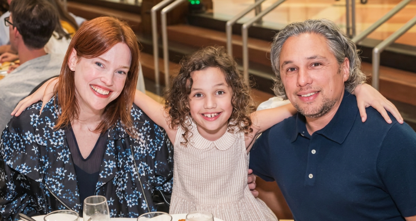 Three people smiling together at a table inside Temple Emanu-El, with a child in the middle warmly embracing two adults.