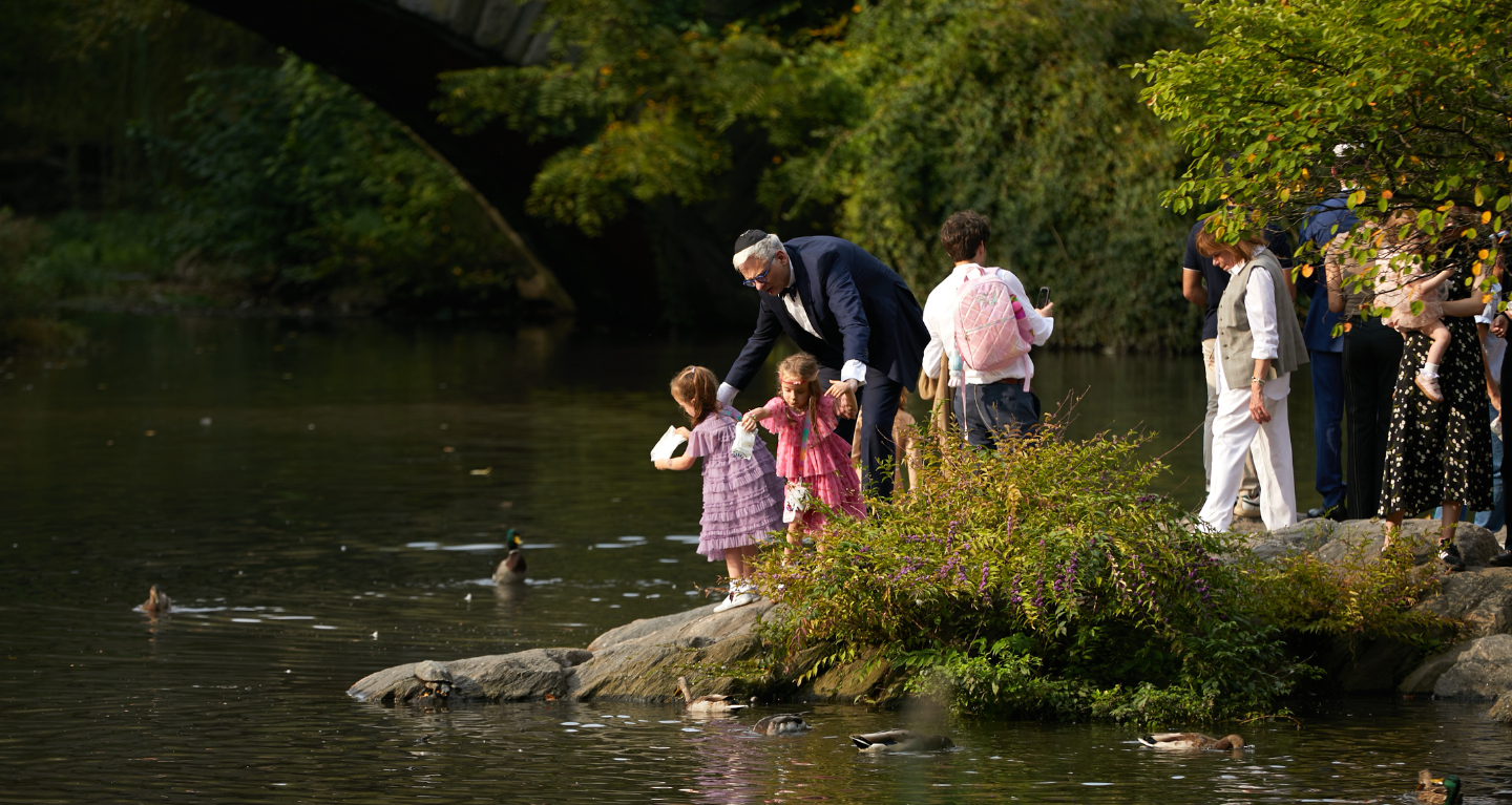 Families, including children, gather beside a pond to feed ducks near a stone bridge in a peaceful park setting.
