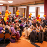 A large indoor gathering at Temple Emanu-El with people seated at round tables, red and yellow balloons decorating the space, and a presentation screen at the front of the room.