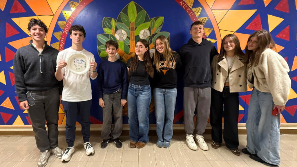 Eight smiling teens stand in front of a vibrant geometric mural featuring a central tree design, gathered at Temple Emanu-El.