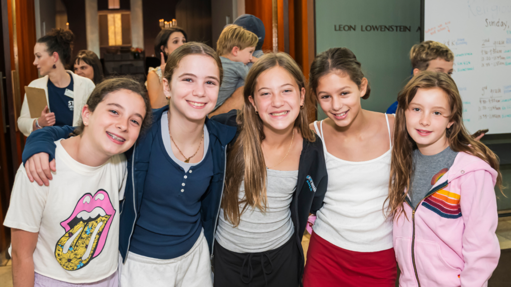 Five smiling girls stand close together indoors at Temple Emanu-El, arms around each other, with other children and adults from the synagogue community in the background.