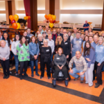 Members and guests gather and smile in a festively decorated Temple Emanu-El hall, surrounded by balloons and tables arranged for a special community celebration.