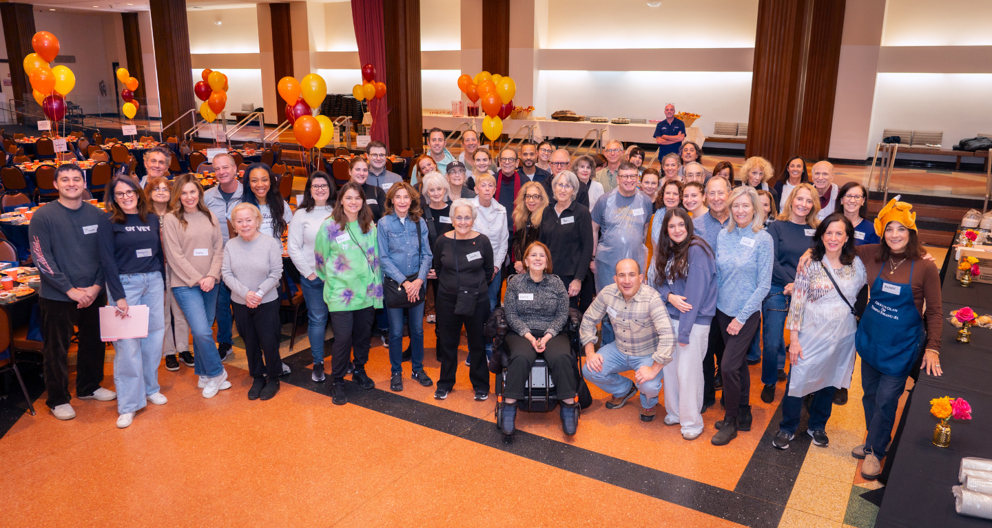 Members and guests gather and smile in a festively decorated Temple Emanu-El hall, surrounded by balloons and tables arranged for a special community celebration.