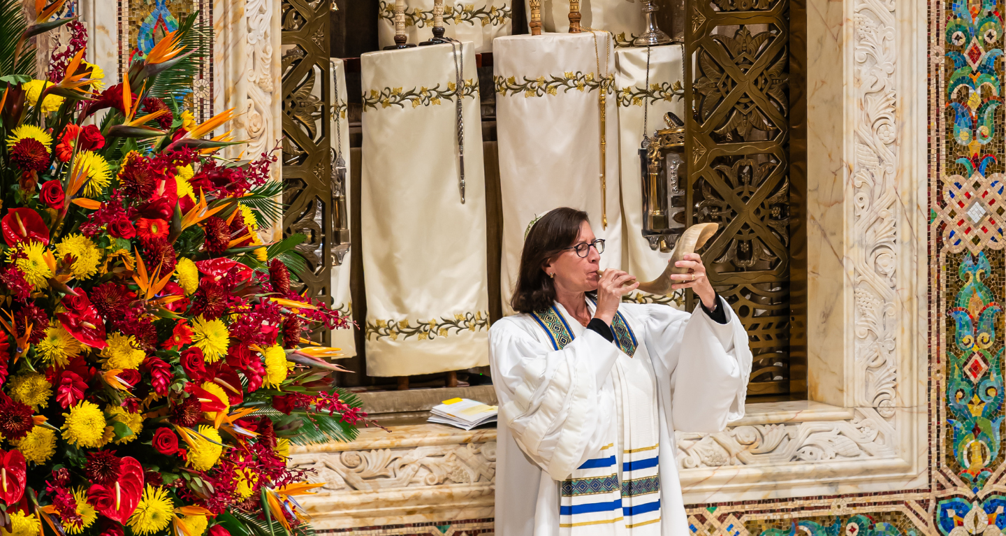 A person in a white robe blows a shofar inside Temple Emanu-El, with vibrant floral arrangements visible on the left.