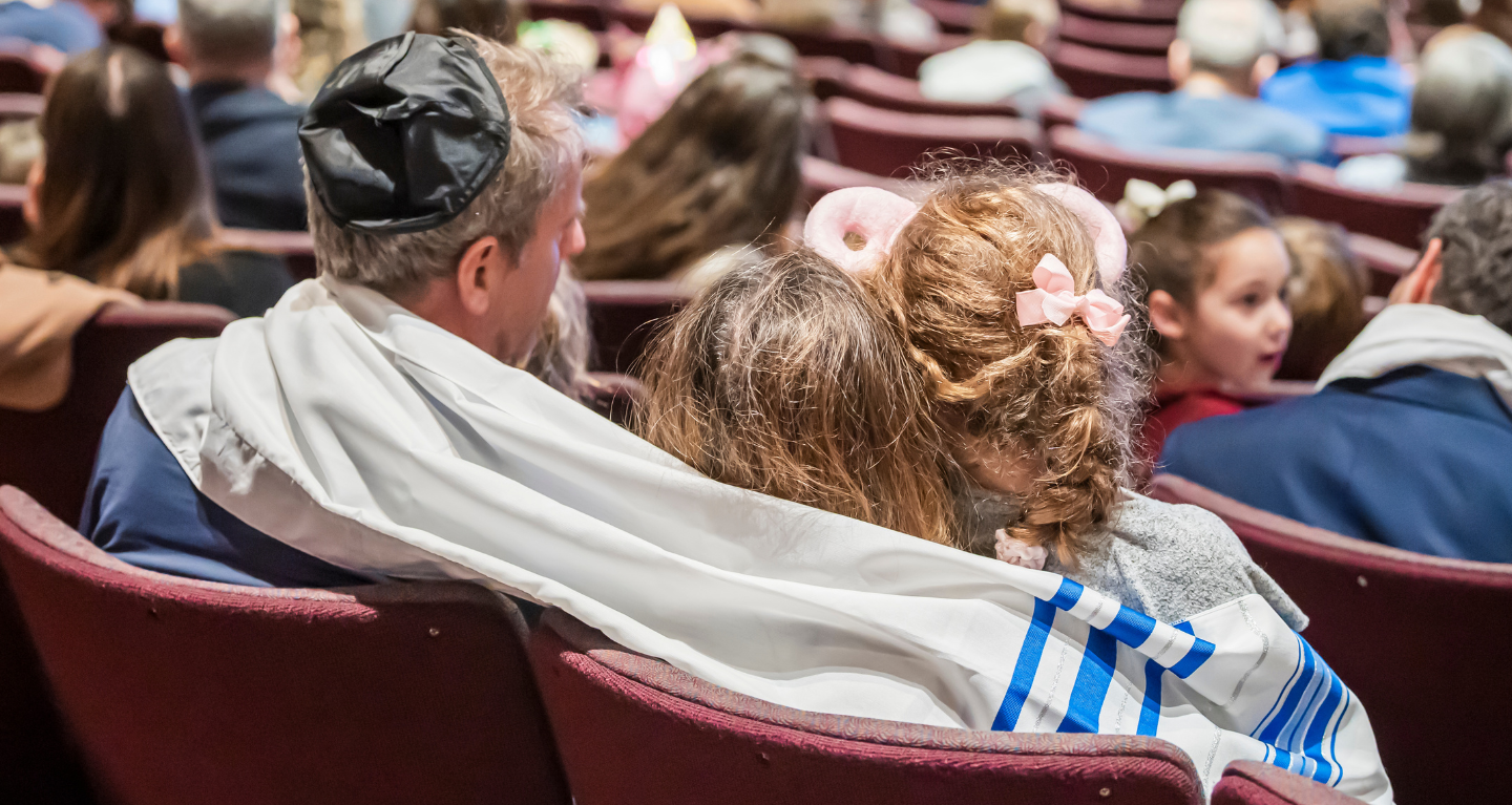 A man wearing a tallit embraces two children while seated among a large gathering in the Temple Emanu-El sanctuary.