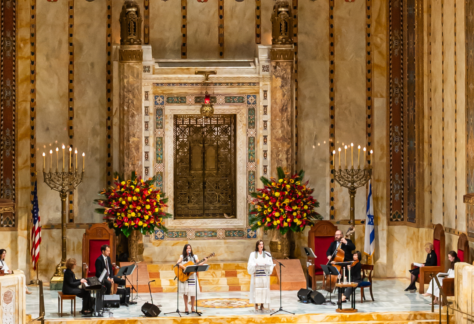 Musicians and vocalists perform in the grand, ornate sanctuary of Temple Emanu-El, with large floral arrangements and decorative elements displayed.