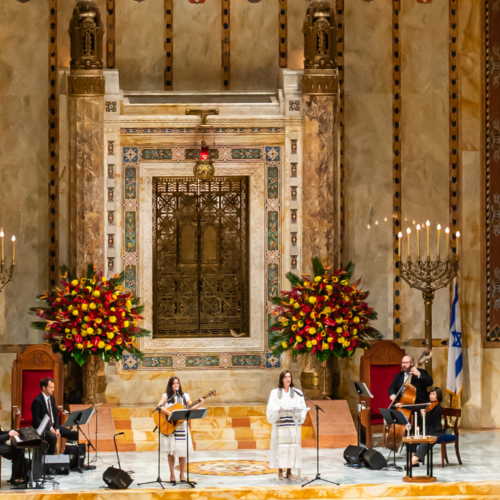 Musicians and vocalists perform in the grand, ornate sanctuary of Temple Emanu-El, with large floral arrangements and decorative elements displayed.
