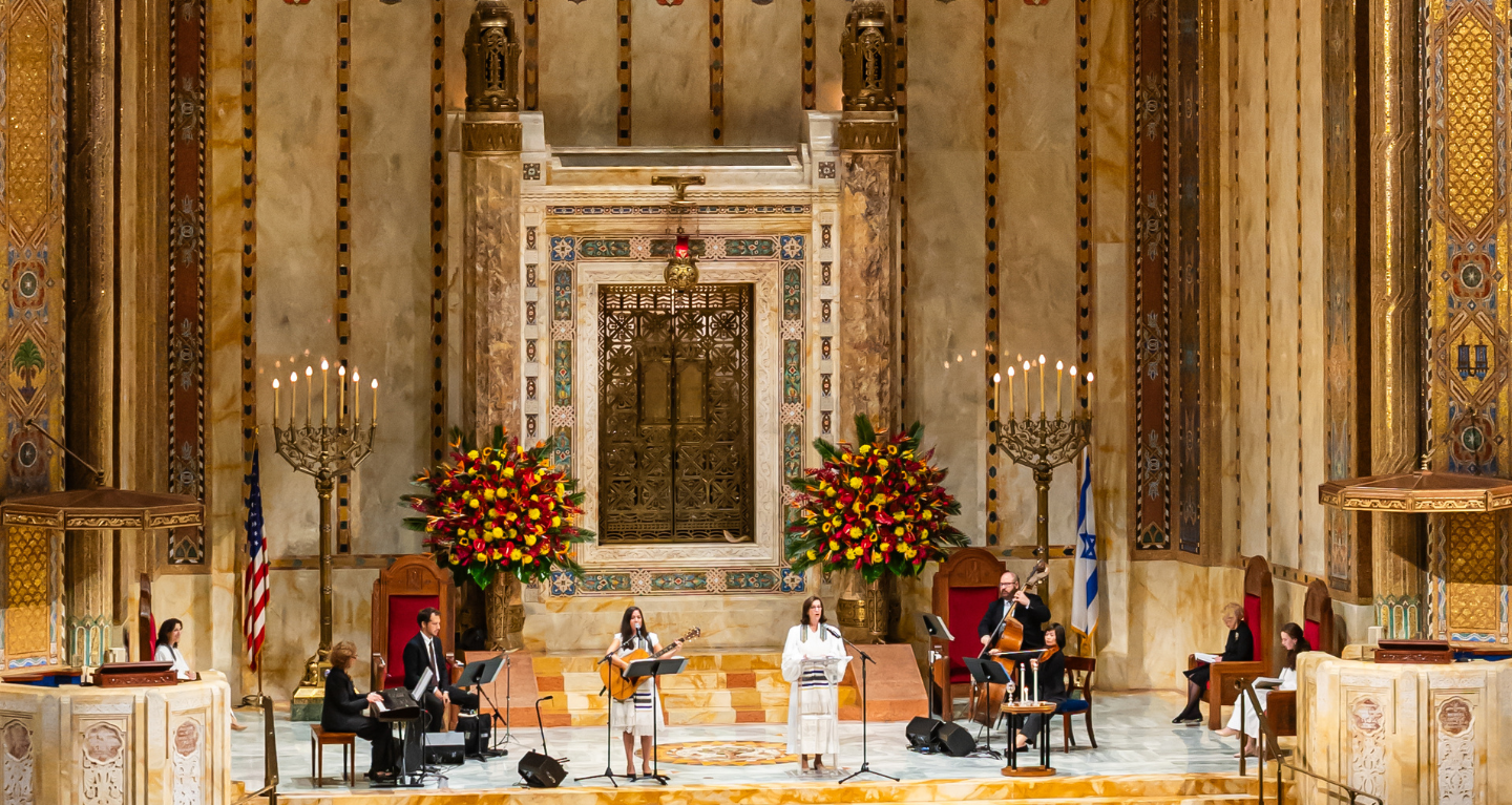 Musicians and vocalists perform in the grand, ornate sanctuary of Temple Emanu-El, with large floral arrangements and decorative elements displayed.