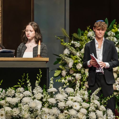 Four young people stand by a podium decorated with flowers in the sanctuary of Temple Emanu-El; two are speaking while the other two hold prayer books and listen attentively.