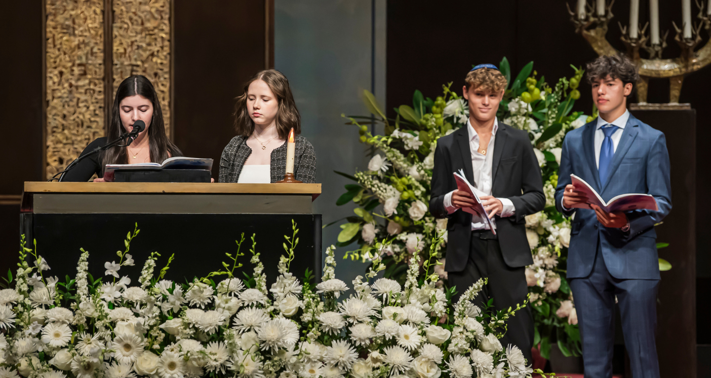 Four young people stand by a podium decorated with flowers in the sanctuary of Temple Emanu-El; two are speaking while the other two hold prayer books and listen attentively.